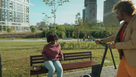 African-American-Man-Riding-E-Scooter-in-Park-and-Chatting-with-Girlfriend-on-Bench